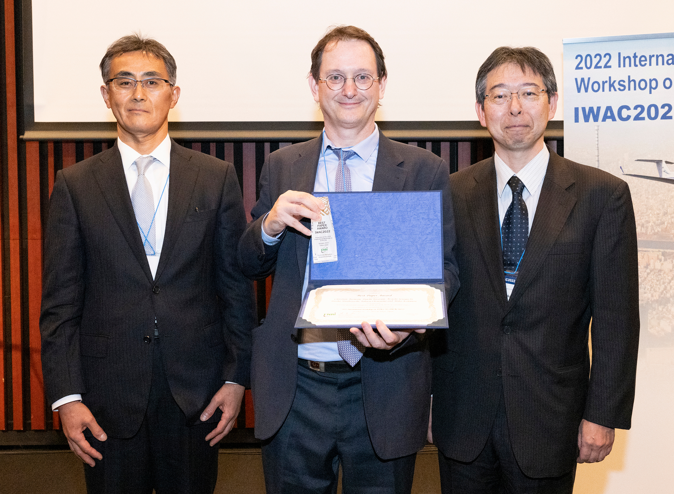 Photo taken at the award ceremony:Three smiling men in suits. Christian Aveneau is in the center, holding an award certificate.