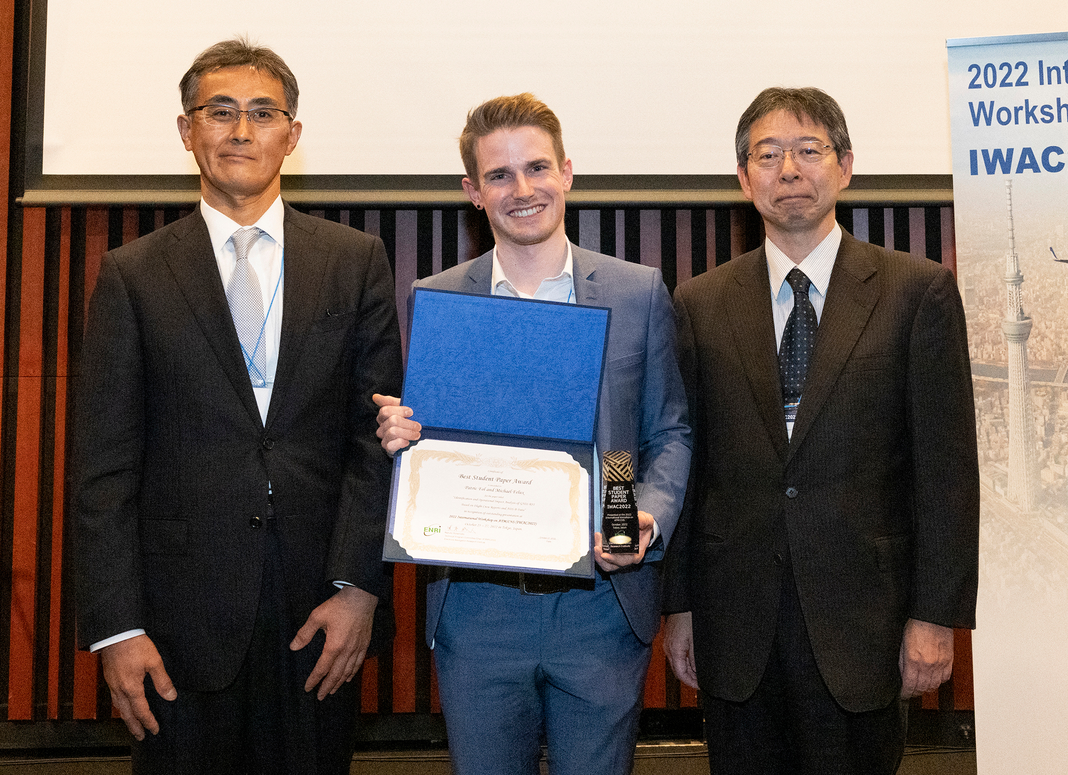 Photo taken at the award ceremony:Three smiling men in suits. Patric Fol is in the center, holding an award certificate.